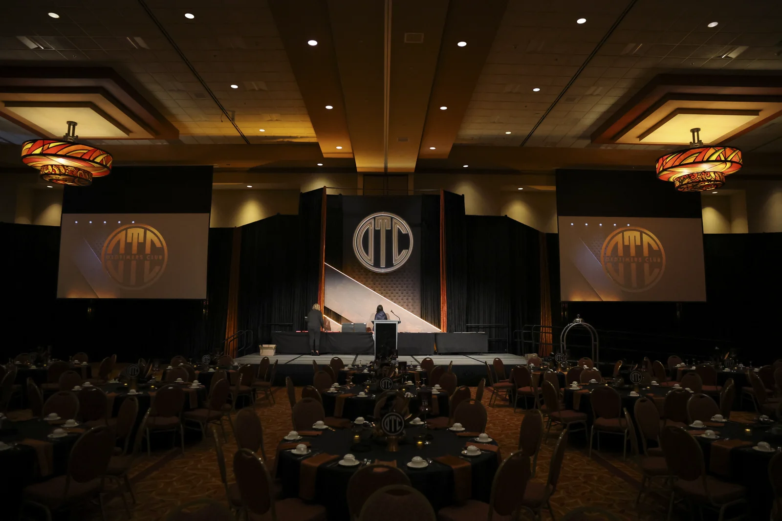Ballroom stage with the illuminated OTC monogram mounted above it, flanked by projection screens