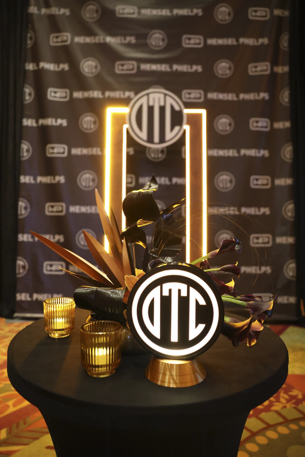 Step-and-repeat backdrop with the Hensel Phelps and OTC marks, an illuminated arch, and a cocktail table