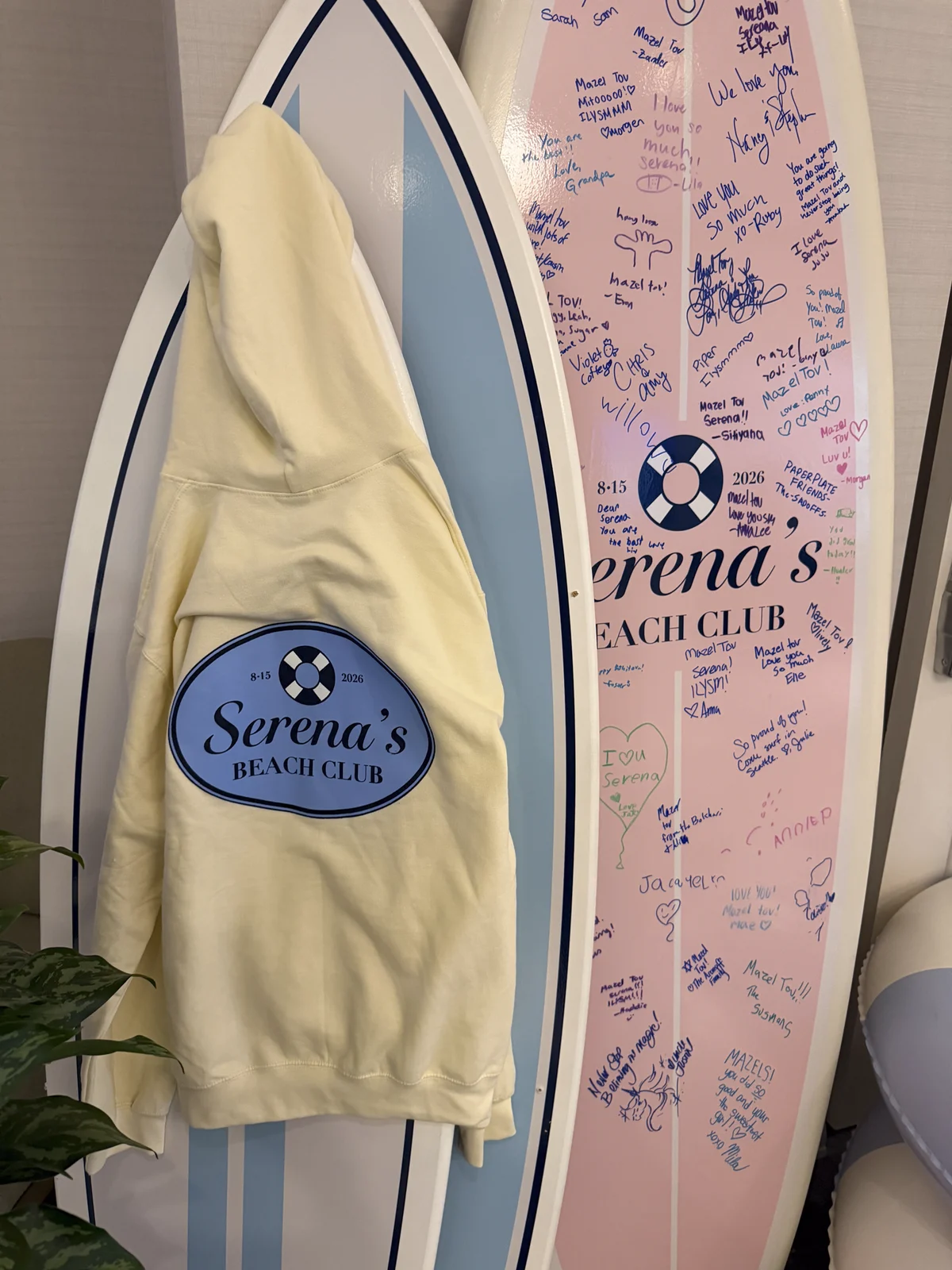 Cream hoodie with the Serena's Beach Club oval patch, hung on the signed surfboard